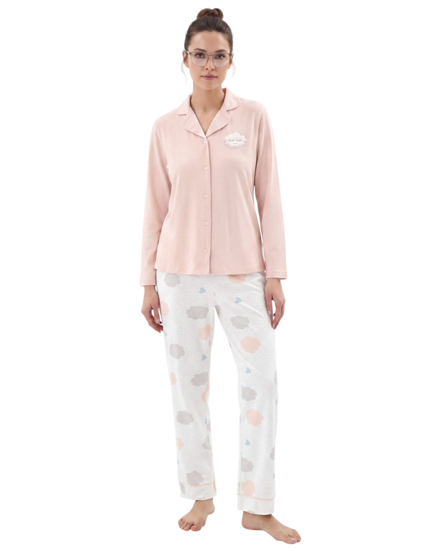 A woman modeling a two-piece pajama set consisting of a light pink long-sleeve button-up top with a small embroidered logo on the left chest, and white pajama pants with a soft pastel cloud pattern in pink, gray