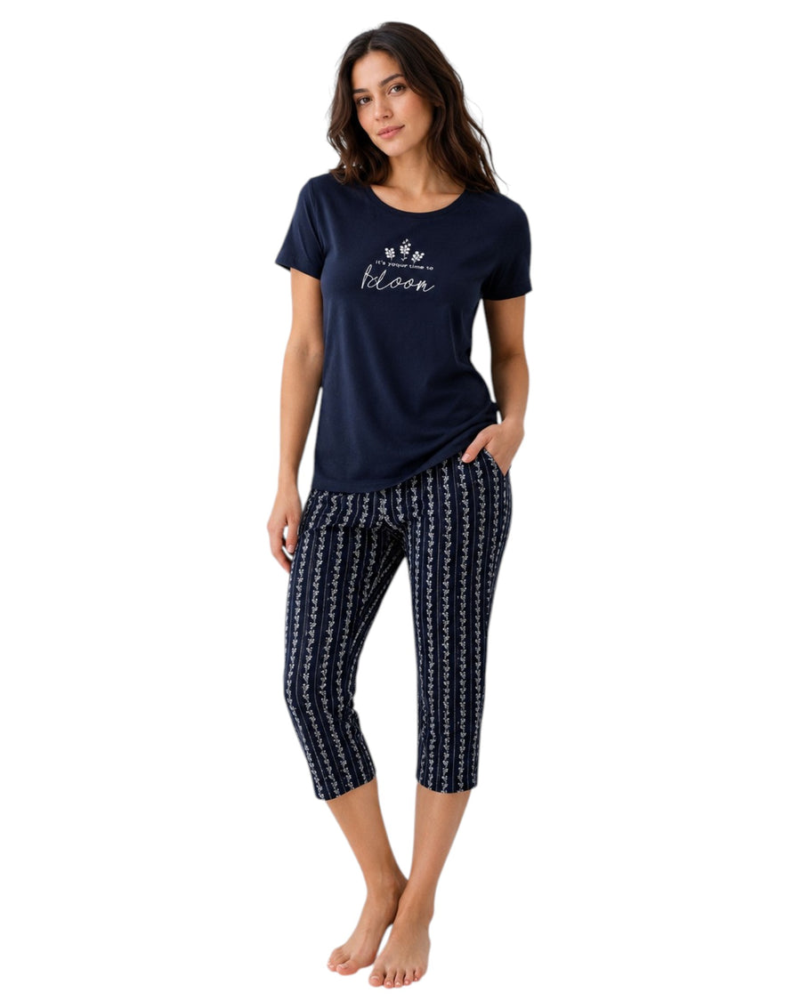 A woman modeling a navy blue two-piece pajama set against a plain white background. The set includes a short-sleeve t-shirt with 'we bloom' printed in white script on the chest, and matching navy capri pants with a white vertical stripe pattern.