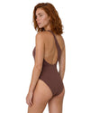 Model wearing a chocolate brown one-shoulder swimsuit with gold-tone ring detail, asymmetric neckline, and open back design.