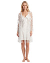 A woman stands barefoot against a plain white studio background wearing a white slip dress under a matching sheer embroidered robe.