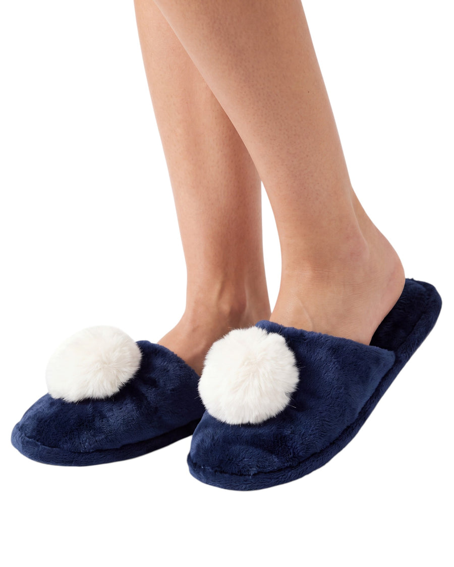 A pair of navy blue plush slip-on slippers with large white pom-poms on the front