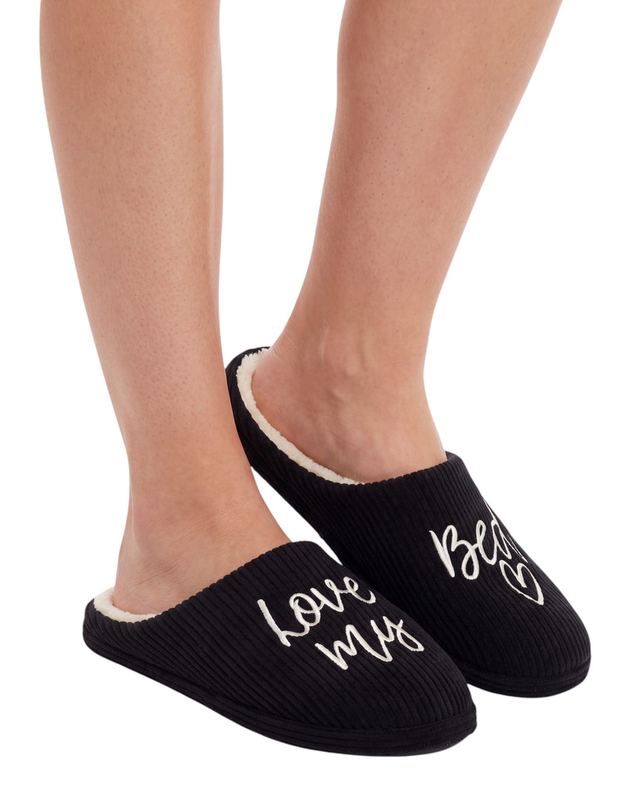 A person wearing black ribbed slip-on slippers with white script text reading 'love my best' and a cream sherpa lining